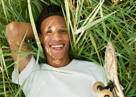 A cheerful young skateboarder lying in the grass on a sunny day, smiling with a skateboard beside him.
